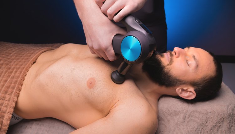 close-up of a professional male masseur massaging pectoral muscles with a percussion instrument, massage gun of a muscular athlete, in a spa treatment, lying on his back on a massage table