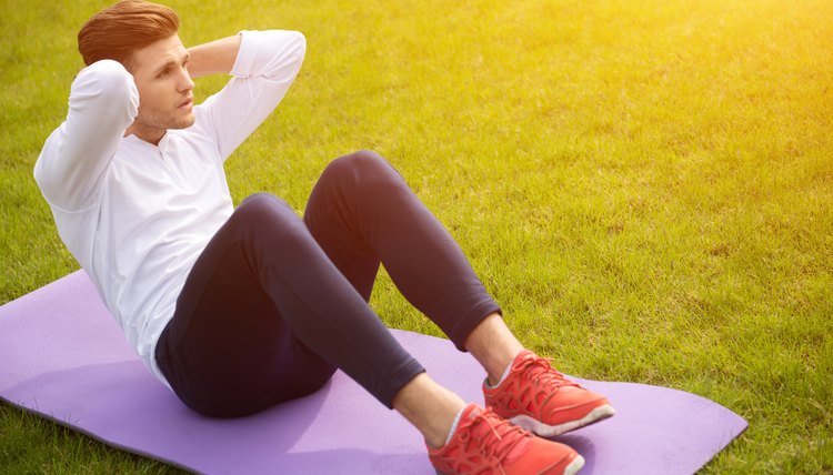 Fit young man doing sit-ups with efforts. He is sitting on mat on grass